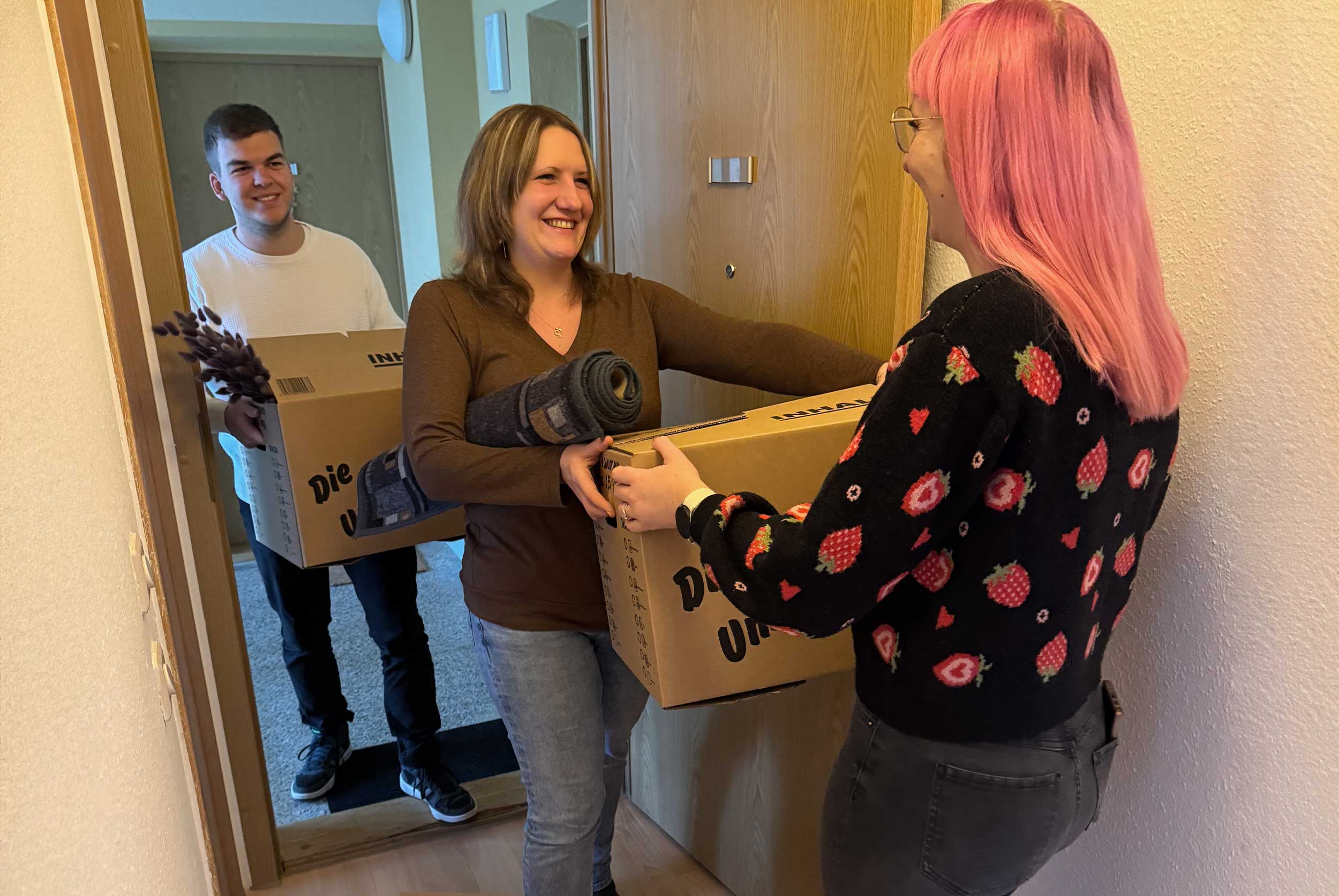 Drei junge Erwachsene stehen an einer Wohnungstür und tragen Umzugskartons in eine Wohnung eines Mehrfamilienhauses.