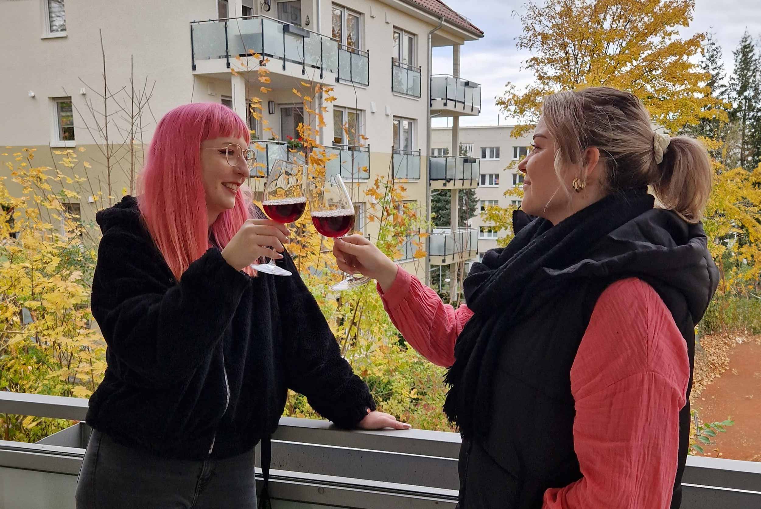 Zwei junge Frauen stoßen mit Weingläsern auf einem Balkon eines Mehrfamilienhauses an.