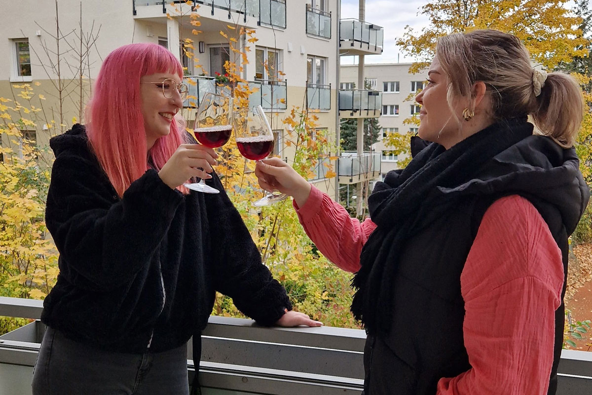 Zwei junge Frauen stoßen mit Weingläsern auf einem Balkon eines Mehrfamilienhauses an.